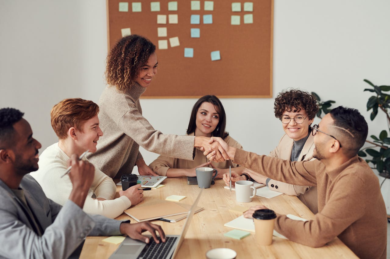 A diverse group of professionals in a modern office setting, engaged in a friendly discussion. A woman in a beige sweater shakes hands with a colleague across the table, symbolizing trust, collaboration, and emotional intelligence in leadership.